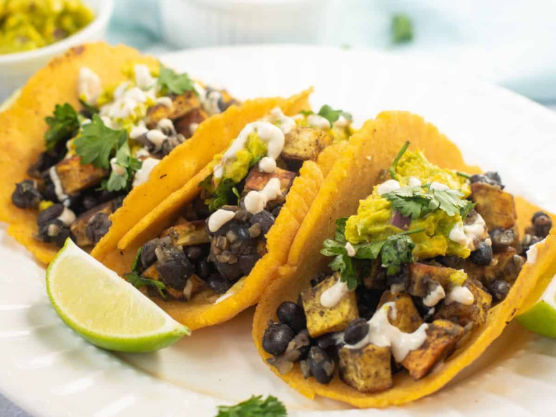 Three tacos filled with black beans, tofu, and vegetables, topped with sauce and cilantro, served with lime wedges on a white plate.