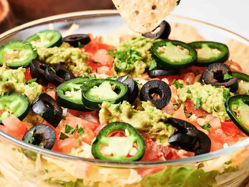 Glass bowl with layered dip topped with guacamole, sliced jalape&ntilde;os, black olives, and tomatoes. A tortilla chip is dipping into the bowl.