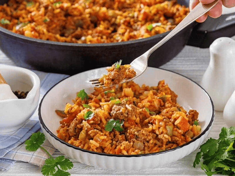 A bowl of Texas hash with rice garnished with cilantro, with a fork lifting a bite, and a skillet of the same dish in the background.
