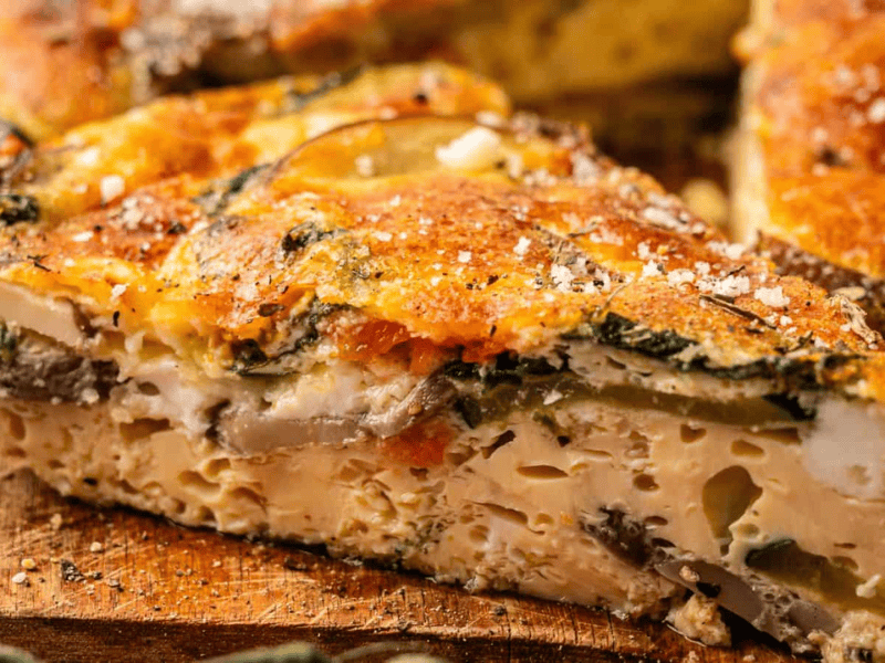 A close-up of a slice of vegetable frittata on a wooden surface, showing layers of egg, vegetables, and seasoning.