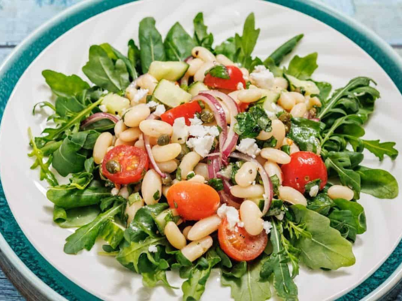 A plate of White Bean Salad with arugula, white beans, cherry tomatoes, red onion, cucumber, and crumbled cheese.