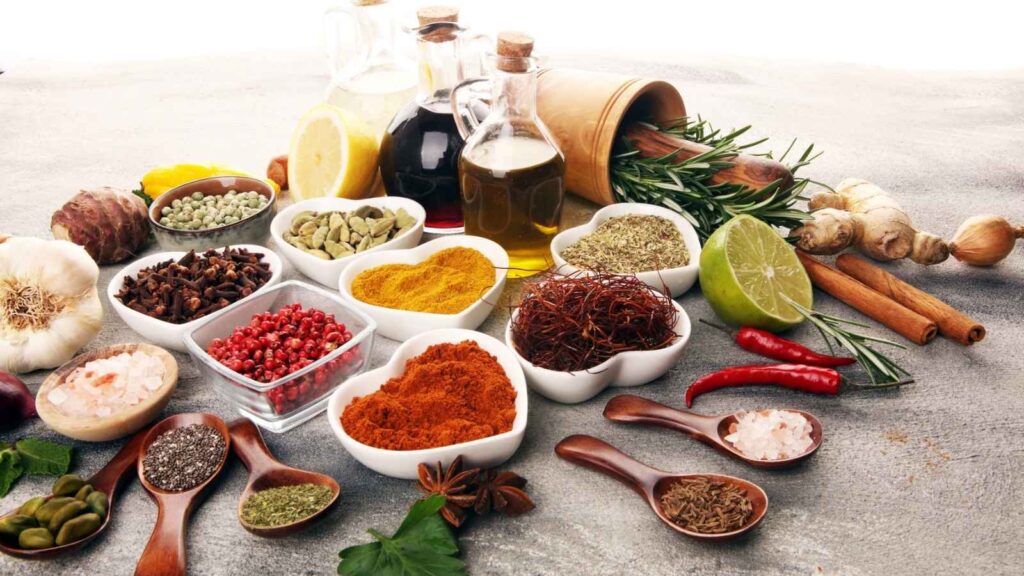 A variety of spices, herbs, and oils arranged in bowls and on a table, including chili peppers, garlic, cinnamon sticks, and glass bottles of oil and vinegar.