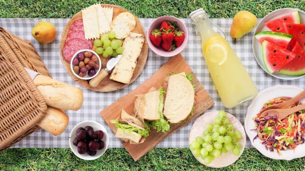 A picnic spread on grass with sandwiches, cheese, crackers, fruit, coleslaw, lemonade, a basket with baguettes, and bowls of grapes, olives, and strawberries on a checkered cloth.