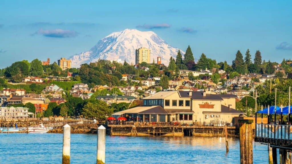 A waterfront building and pier in the foreground, with a residential neighborhood and a snow-capped mountain in the background under a clear blue sky.