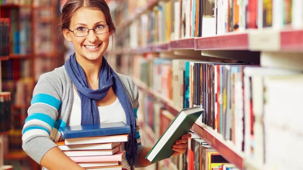 Woman wearing glasses and a scarf stands in a library aisle, smiling while holding a stack of books in one arm and another book in her other hand.