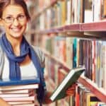 Woman wearing glasses and a scarf stands in a library aisle, smiling while holding a stack of books in one arm and another book in her other hand.