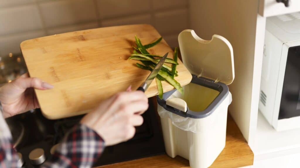 A person scrapes vegetable peels off a wooden cutting board into a kitchen trash bin, considering apartment composting as an eco-friendly alternative.