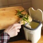 A person scrapes vegetable peels off a wooden cutting board into a kitchen trash bin, considering apartment composting as an eco-friendly alternative.