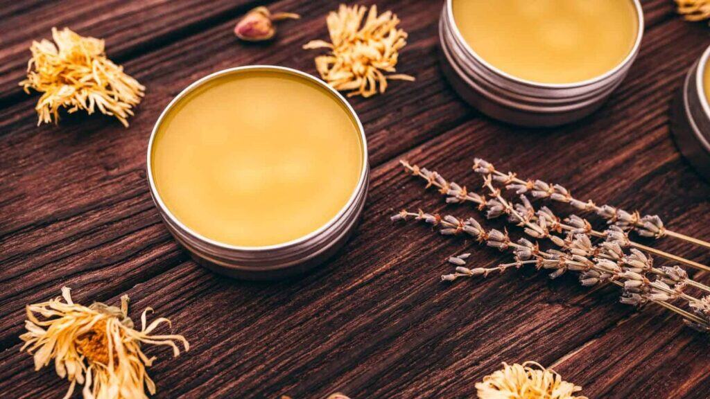 Two metal tins filled with yellow balm sit on a wooden surface, surrounded by dried flowers and lavender sprigs—a perfect scene for DIY beauty enthusiasts.