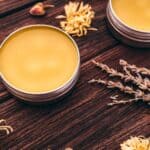 Two metal tins filled with yellow balm sit on a wooden surface, surrounded by dried flowers and lavender sprigs&mdash;a perfect scene for DIY beauty enthusiasts.