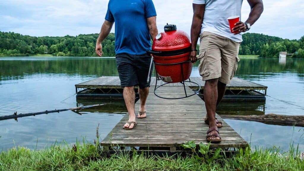 Two men walk on a dock by a lake, carrying a red portable grill and a red cup&mdash;creating the perfect outdoor kitchen scene amid trees and sparkling water.