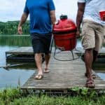 Two men walk on a dock by a lake, carrying a red portable grill and a red cup—creating the perfect outdoor kitchen scene amid trees and sparkling water.