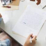 A person with curly hair writes a letter at a white table with a cup of tea, chocolate, and photographs nearby.