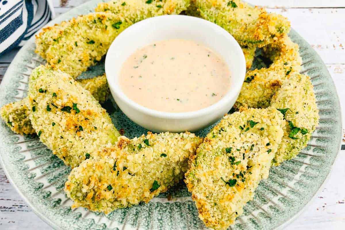 A plate of breaded avocado slices arranged around a bowl of creamy dipping sauce on a green-rimmed dish.