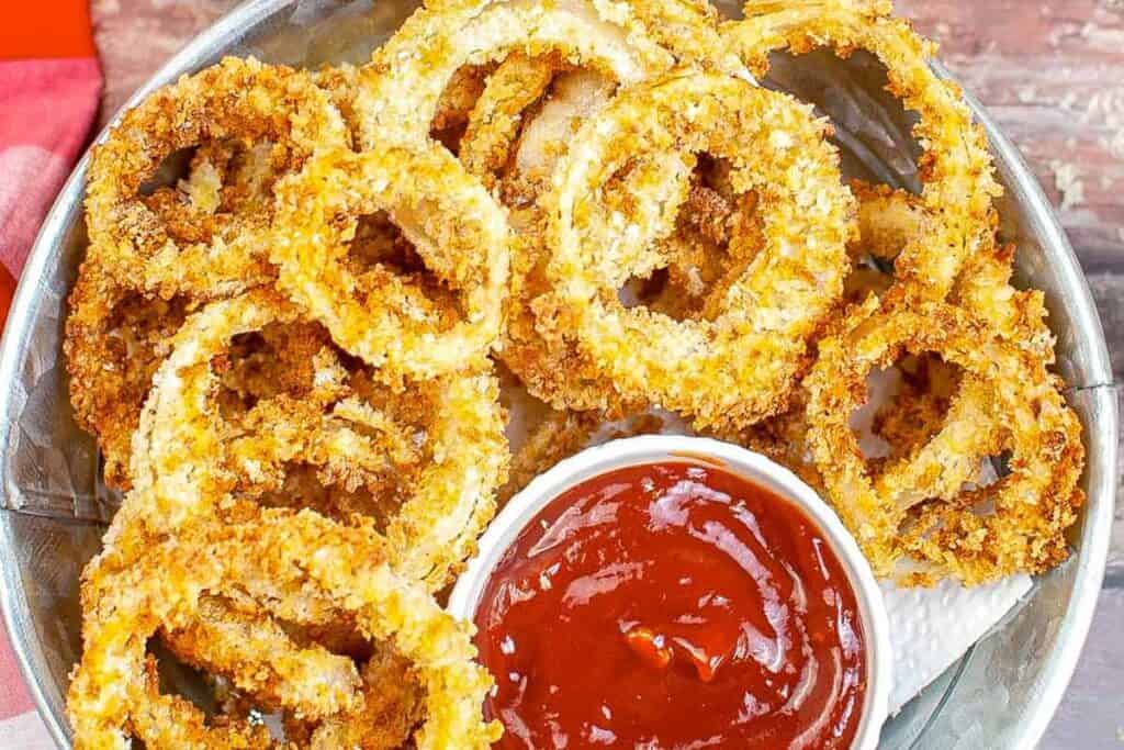 A bowl of crispy onion rings with a small dish of ketchup for dipping on the side.