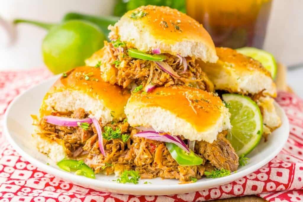 Three pulled pork sliders with shredded meat, sliced onions, jalapenos, and parsley on a white plate, garnished with lime wedges.