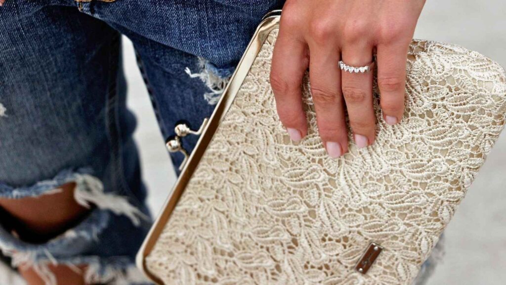 A hand with a diamond ring rests on a cream-colored lace clutch purse; the person is wearing distressed denim jeans.