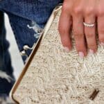 A hand with a diamond ring rests on a cream-colored lace clutch purse; the person is wearing distressed denim jeans.