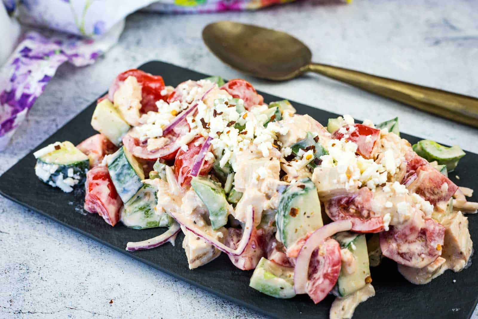 A plate of Greek Salad with Chicken sits on a dark serving tray next to a spoon.