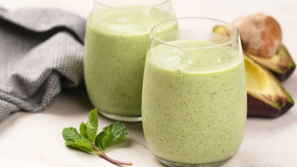Two glasses of green smoothie sit on a light surface next to a sprig of mint, a gray cloth, and a halved avocado with the pit.