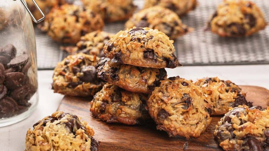 A pile of coconut and chocolate chip cookies on a wooden board, with more cookies cooling on a wire rack in the background.