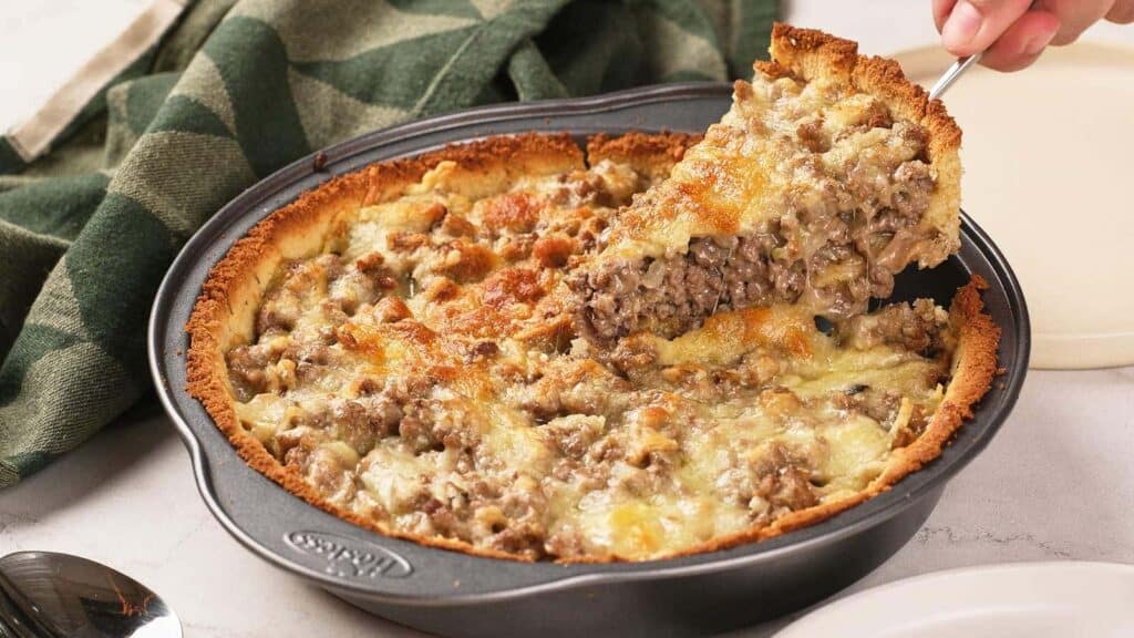 A hand lifts a slice of savory pie from a round baking dish filled with a ground meat and cheese mixture, with a browned crust and a green towel in the background.