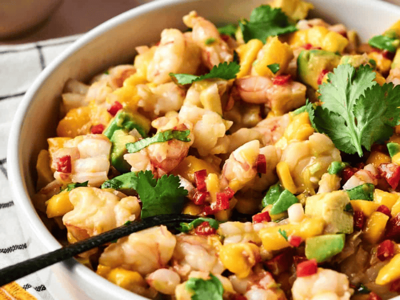 A bowl of shrimp ceviche with diced mango, avocado, red onion, red pepper, and cilantro, with a spoon inside.