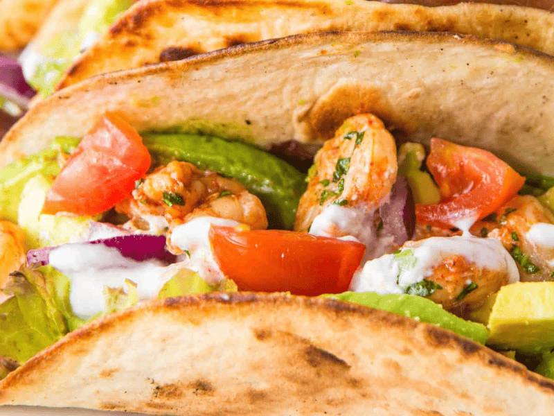 Close-up of a shrimp taco with avocado, tomato, red onion, and a drizzle of sauce in a toasted tortilla.