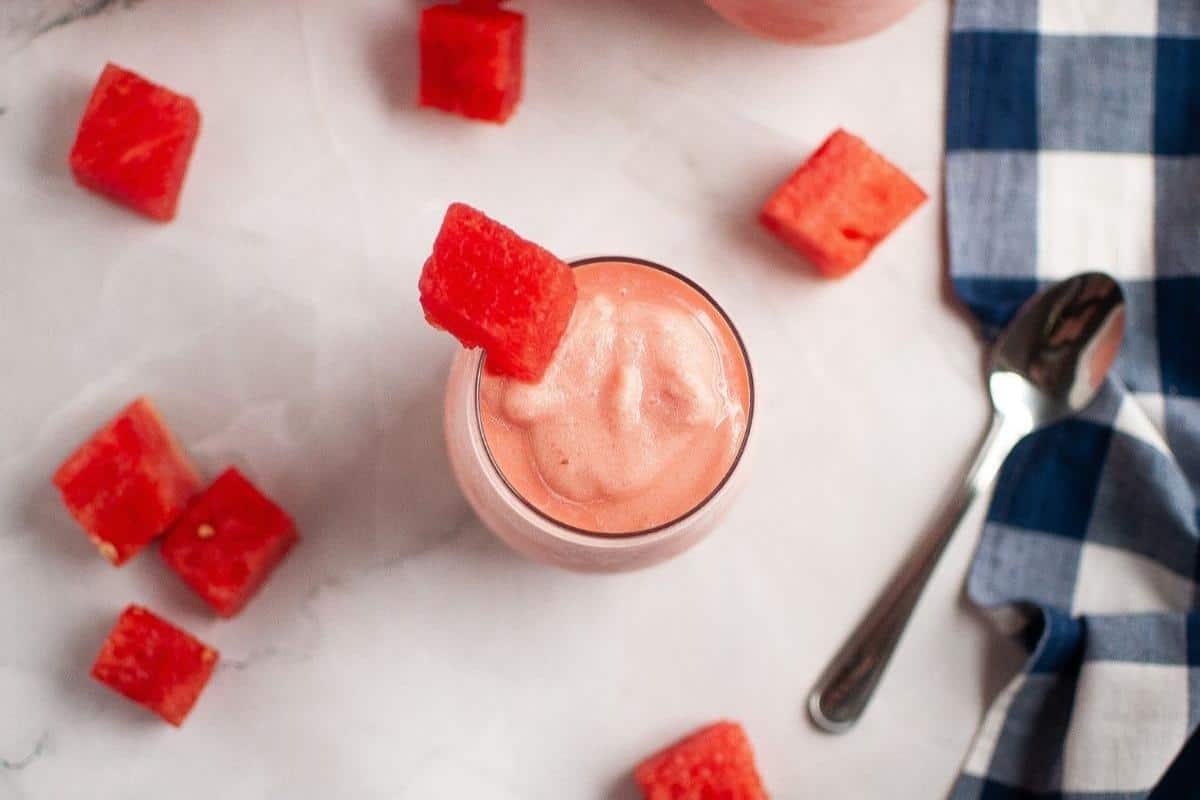 A glass of pink watermelon smoothie garnished with a watermelon cube, surrounded by watermelon pieces, a spoon, and a blue checkered cloth on a marble surface.