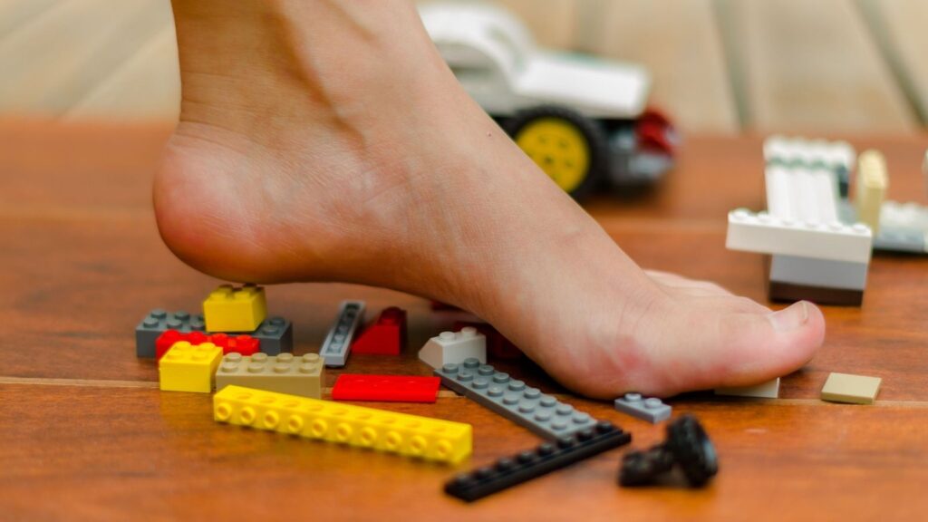 A bare foot stepping on scattered Lego bricks on a wooden floor with more Lego pieces and a toy car in the background.