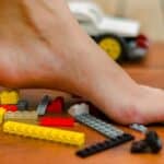 A bare foot stepping on scattered Lego bricks on a wooden floor with more Lego pieces and a toy car in the background.