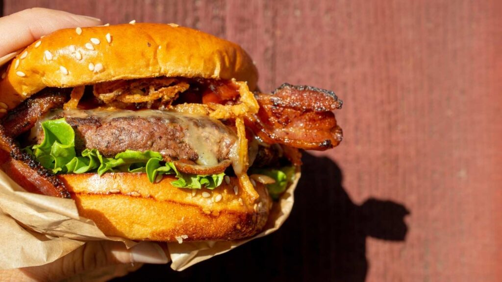 A hand holds a cheeseburger with bacon, crispy onions, lettuce, and a sesame seed bun against a red wooden background.