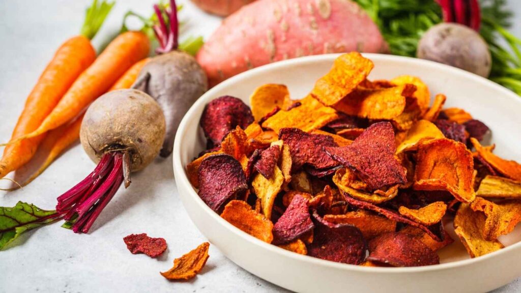 A bowl of colorful vegetable chips with fresh carrots, beets, and a sweet potato on a white surface.