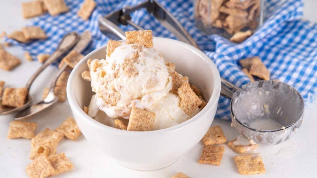 A bowl of vanilla ice cream topped with square cereal pieces sits on a white surface, surrounded by more cereal, an ice cream scooper, and a blue checkered cloth.