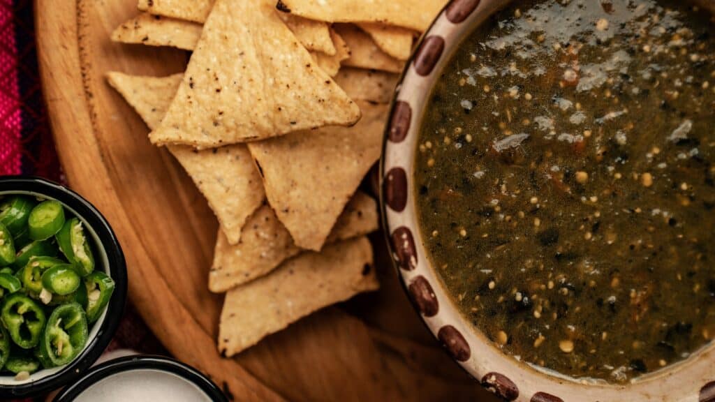 A plate of tortilla chips sits next to a bowl of dark green salsa, with a small dish of sliced green peppers nearby—perfect for celebrating National Salsa Month.
