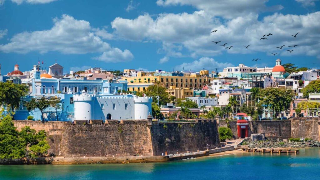 Colorful buildings and a historic fort overlook the blue water in Old San Juan, Puerto Rico, under a partly cloudy sky with birds flying overhead.