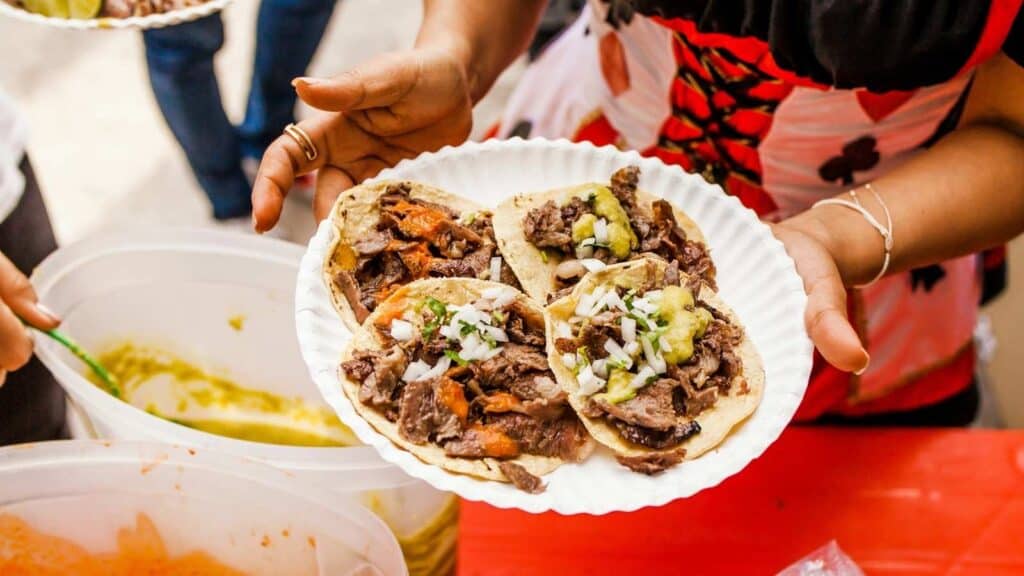 Person holding a paper plate with four beef tacos topped with chopped onions and green salsa, standing next to containers of food.