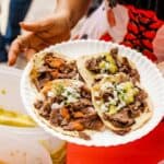 Person holding a paper plate with four beef tacos topped with chopped onions and green salsa, standing next to containers of food.