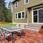 Outdoor patio with a glass-top table and four chairs on a brick surface, next to a brown house with sliding glass doors and steps, overlooking a grassy yard and trees.