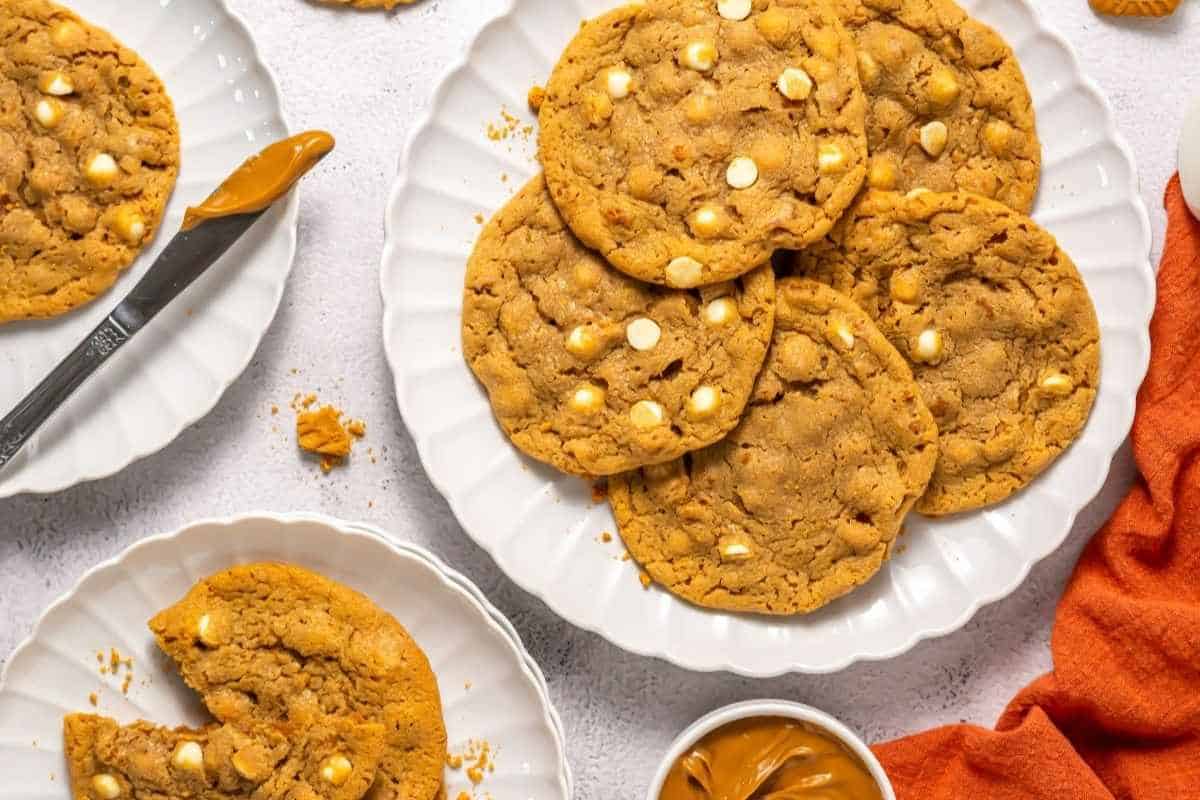 A plate of large peanut Copycat Crumbl Biscoff cookies with white chocolate chips, surrounded by a knife, a small bowl of peanut butter, and an orange napkin.