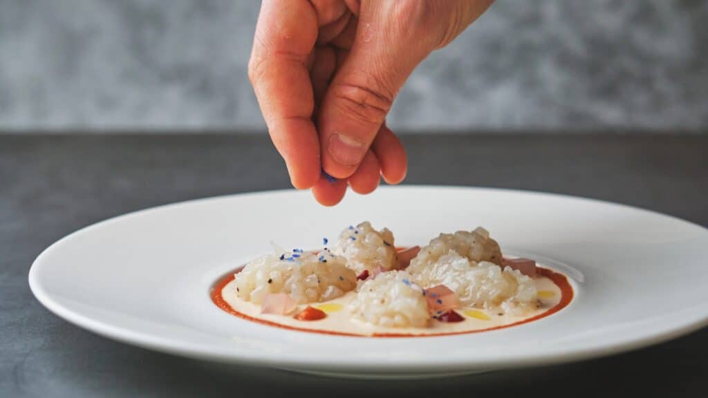 A hand sprinkles seasoning onto a white plate with plated risotto-style rice and sauce, arranged in small portions.