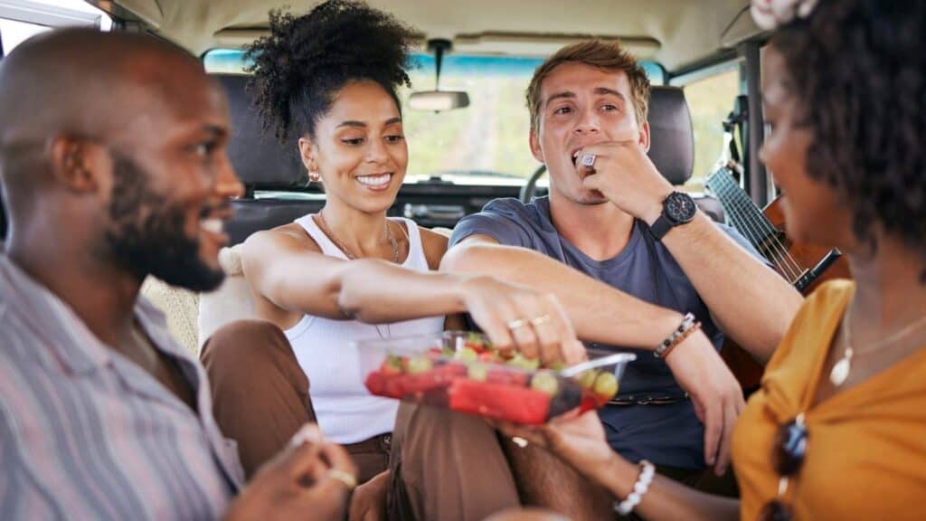 Four people sit inside a vehicle on Memorial Day weekend, sharing and eating a tray of mixed fruit, smiling and enjoying each other's company.