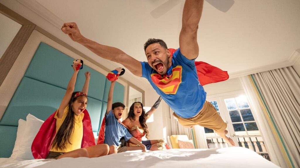 A man in a superhero costume jumps onto a bed while three children in costumes cheer and watch from the bed in a brightly lit bedroom.
