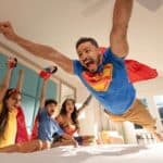 A man in a superhero costume jumps onto a bed while three children in costumes cheer and watch from the bed in a brightly lit bedroom.