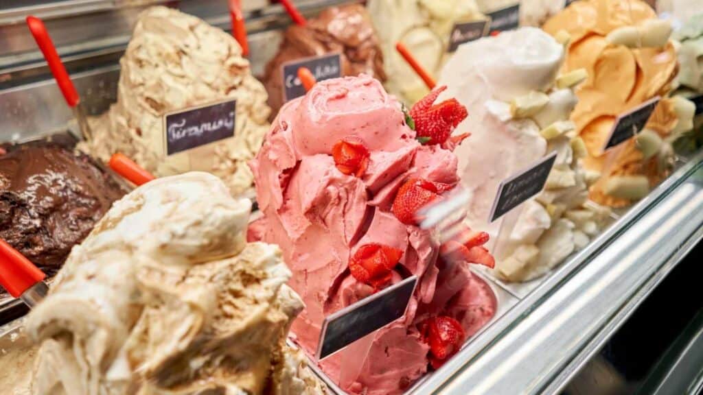 Various flavors of gelato are displayed in a shop, with colorful scoops arranged in tall mounds and small signs indicating their names.