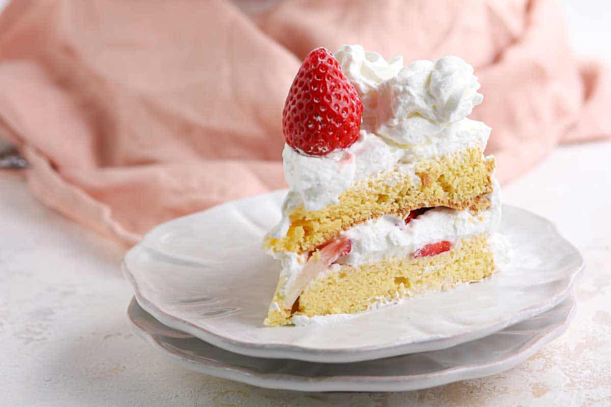 A slice of strawberry shortcake with whipped cream on a white plate, topped with a whole strawberry.