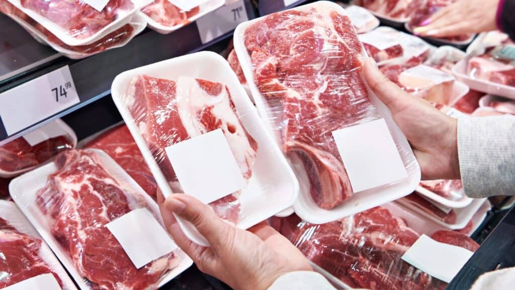 Person holding two packs of raw meat in plastic trays with blank labels at a supermarket meat section.