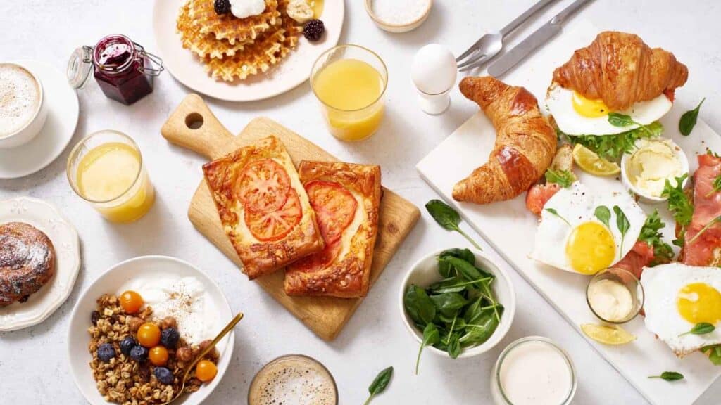 A delightful Mother&rsquo;s Day brunch spread with croissants, fried eggs, smoked salmon, tomato pastries, granola with fruit, waffles, orange juice, coffee, and small bowls of spinach and yogurt.