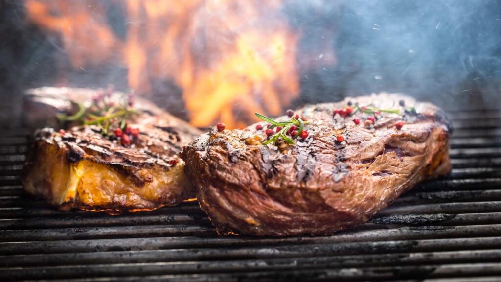 Two thick steaks with herbs and pepper on top are being grilled over open flames, with smoke rising in the background.