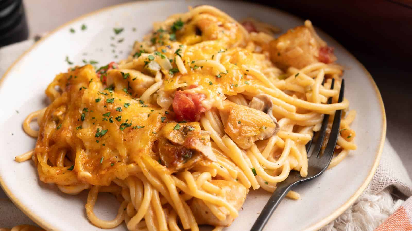 A plate of creamy baked spaghetti with chunks of chicken, diced tomatoes, melted cheese, and herbs, served with a black fork.